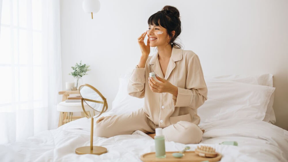 Smiling woman applying face cream sitting on bed